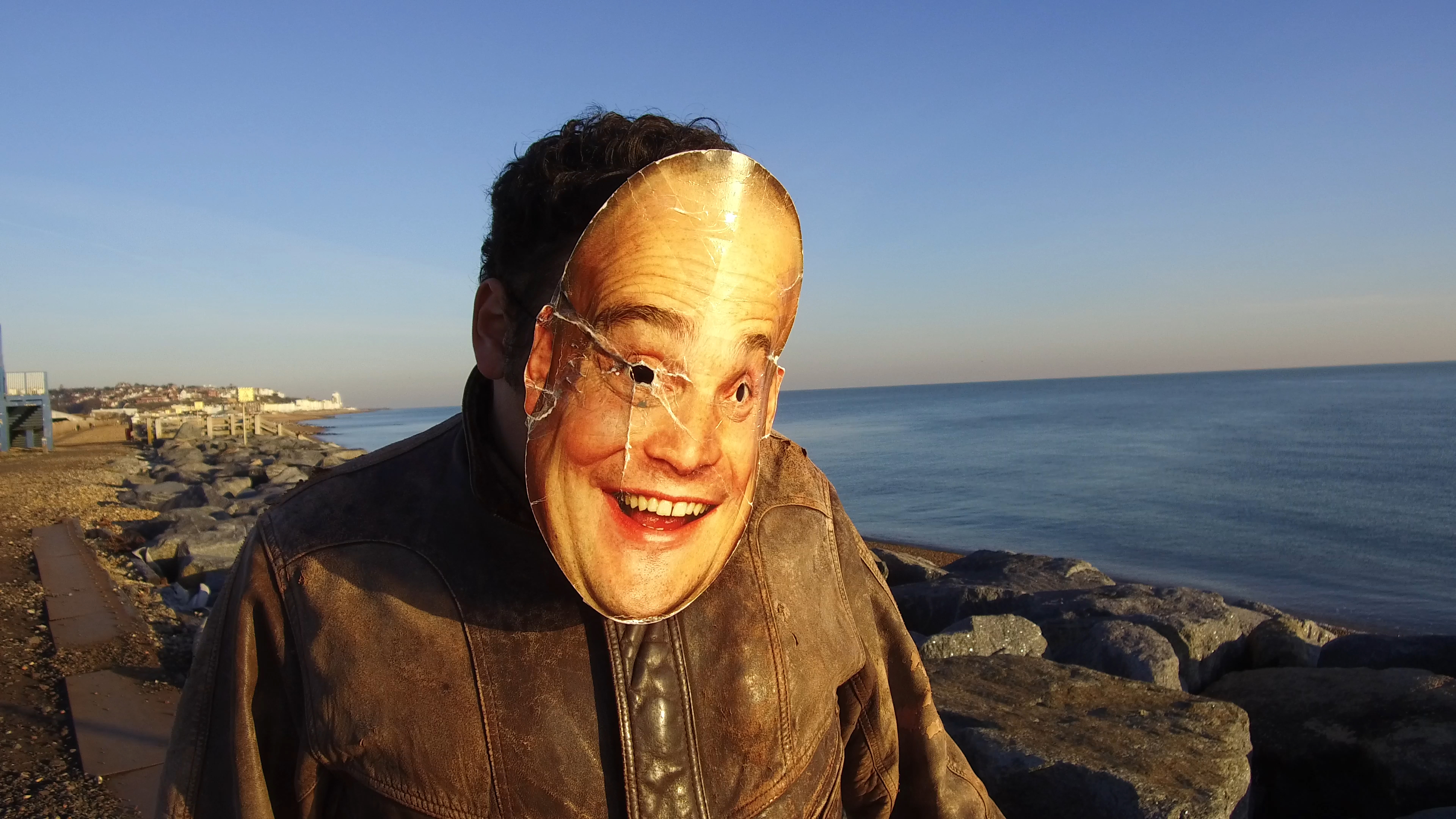 Person wearing a mask of Al Murray, standing on a seafront